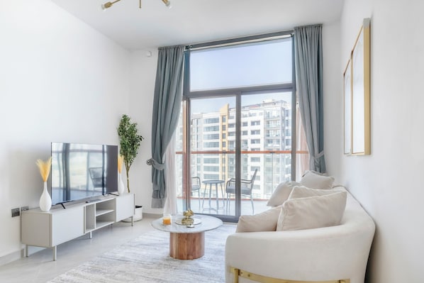 A modern living room with a flat-screen TV, a sofa, a coffee table, and a view of a cityscape through large windows. - Bloomfields 1BR in Binghatti Avenue-Jaddaf (Dubai)