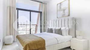 A bedroom with a large window, a bed with a white headboard, a bedside table with a lamp, and a framed picture on the wall. - 122 Bloomfields Cozy1BR Binghatti Avenue (Dubai)