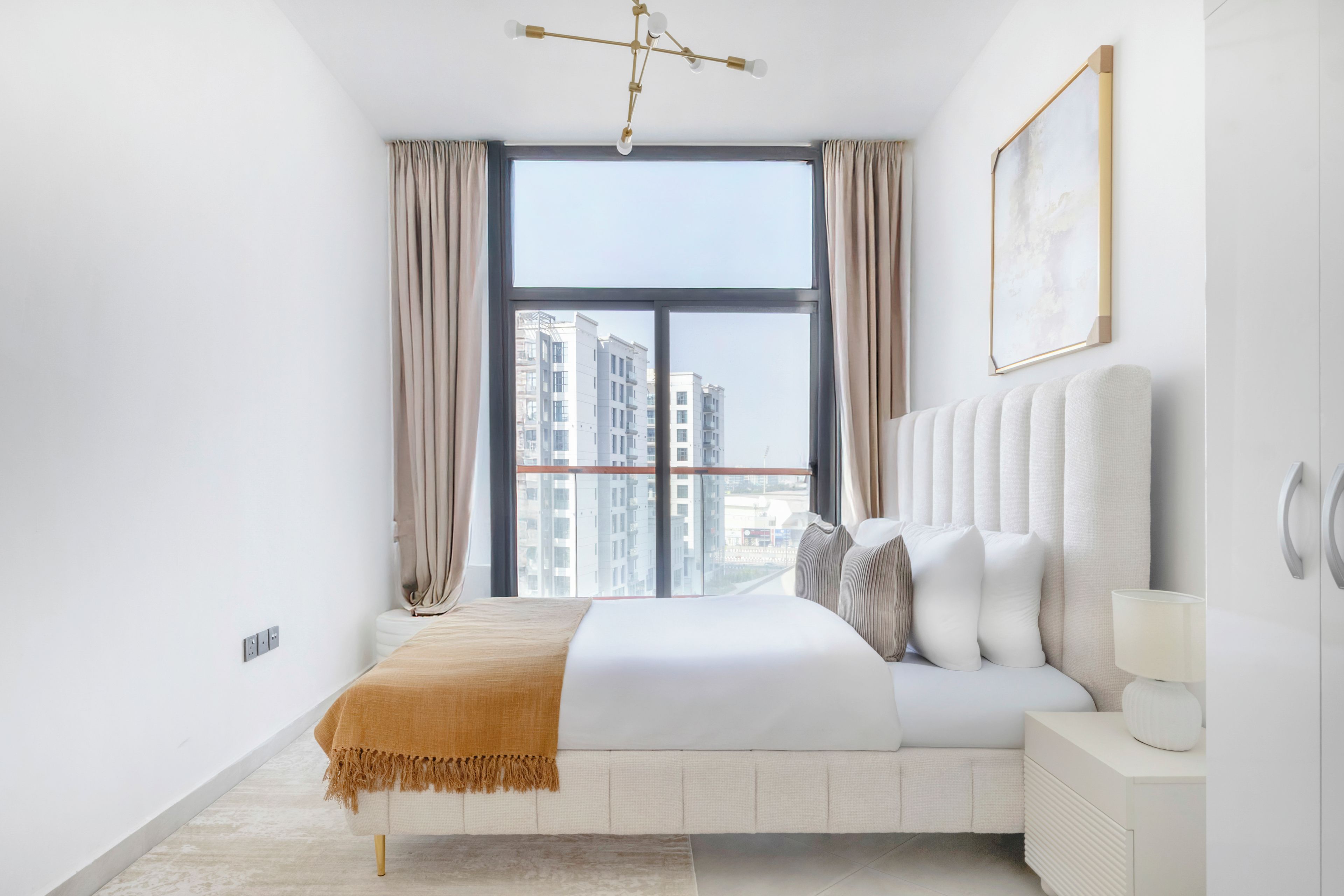 Une chambre moderne avec une grande fenêtre, un lit recouvert de linge de lit blanc, une table de chevet avec une lampe, et une vue sur des immeubles de grande hauteur.