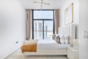 A modern bedroom with a large window, a bed with white bedding, a bedside table with a lamp, and a view of high-rise buildings.