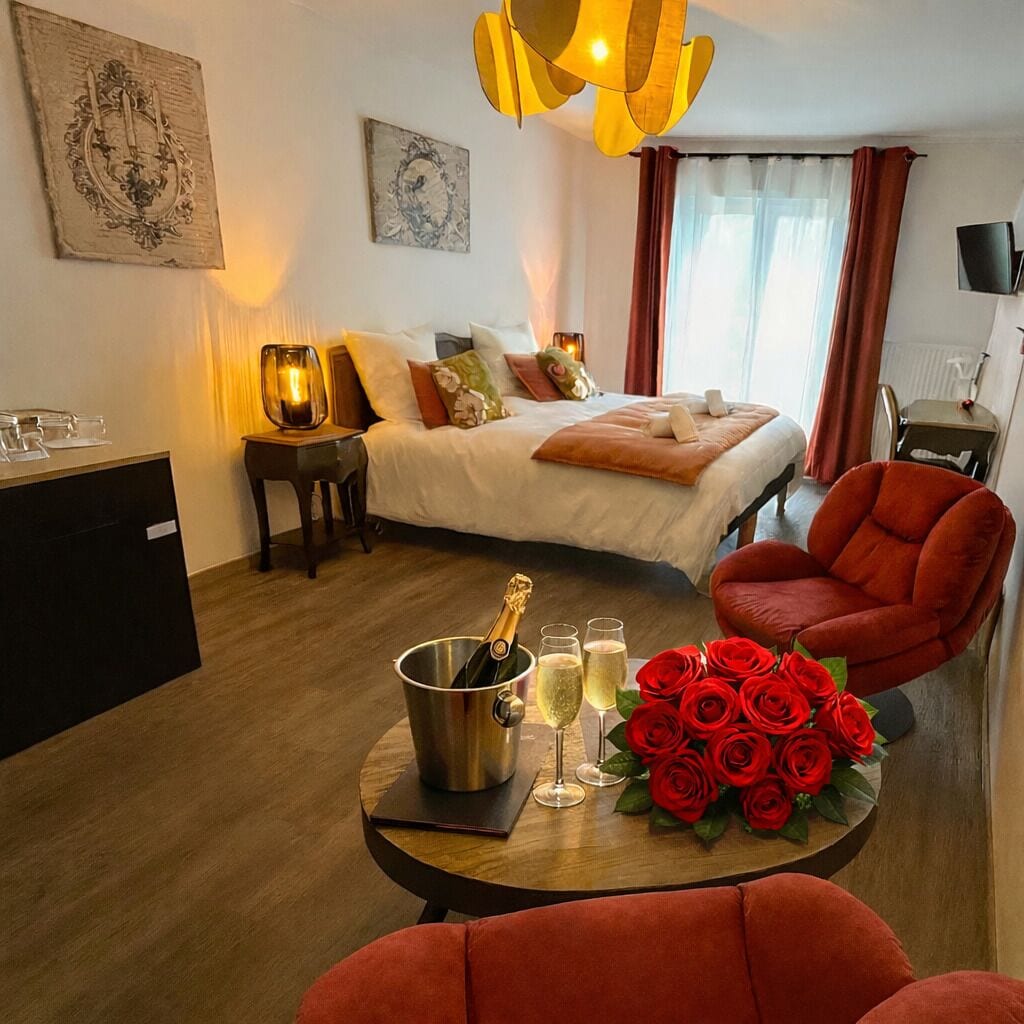Une chambre d’hôtel avec un lit, un bureau, une chaise, une table sur laquelle se trouve une bouteille de champagne, des verres et un bouquet de roses rouges.