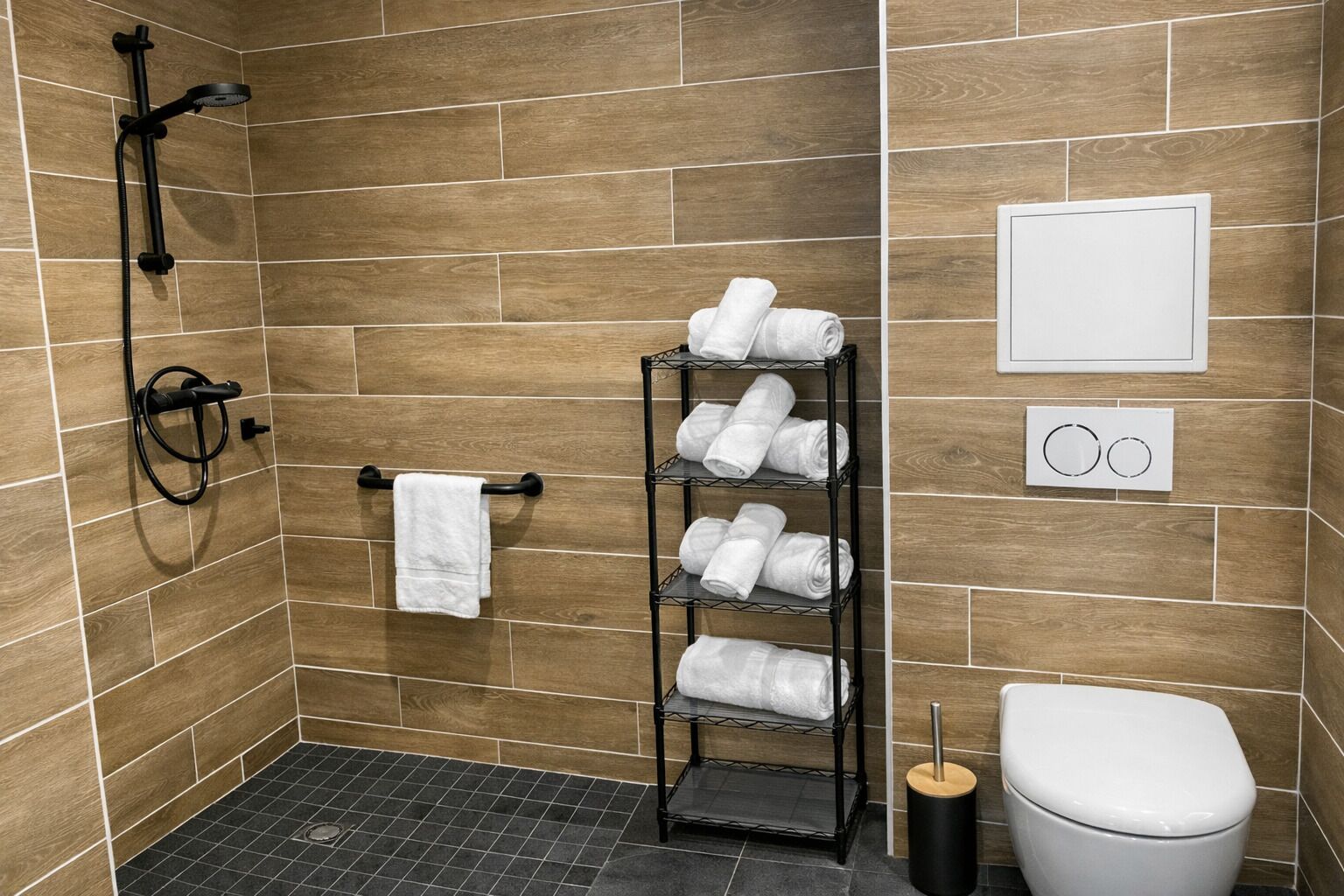 A shower area with a black hose, a towel rack with a white towel, a metal shelf with folded towels, a toilet, and a wall-mounted flush handle.
