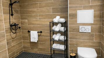 A shower area with a black hose, a towel rack with a white towel, a metal shelf with folded towels, a toilet, and a wall-mounted flush handle.