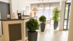 A reception area with a counter, potted plants, a chair, and a chandelier. - Logis Hôtel Blue Garden (Grandvillers)