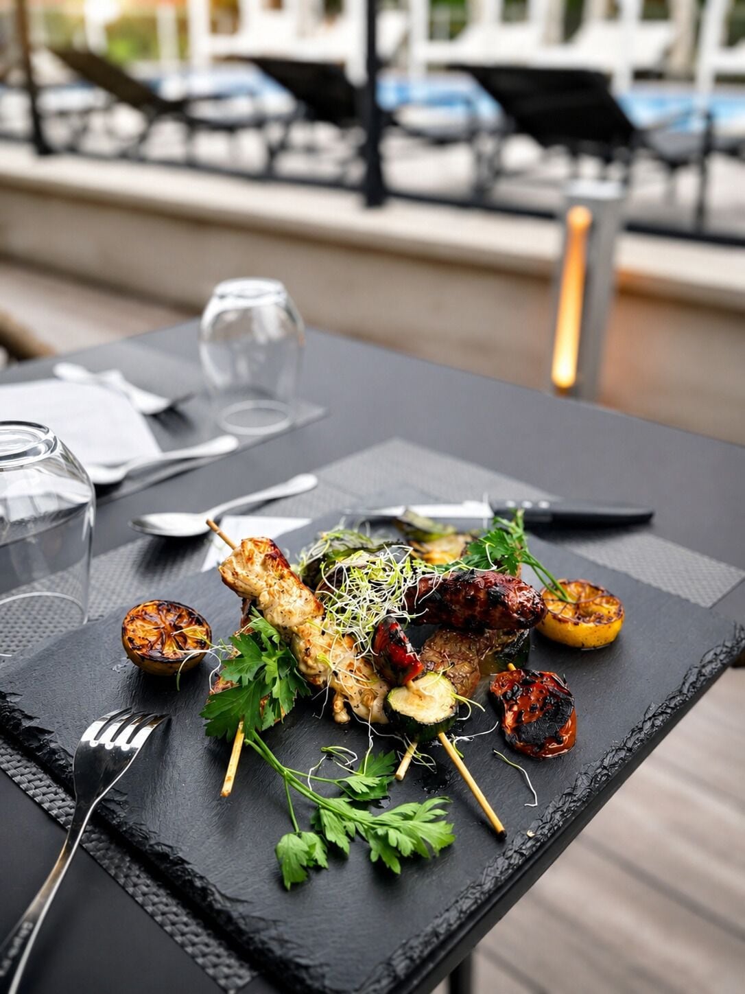 A dining table with a black slate top, a glass of water, a fork, and a plate with grilled skewers and vegetables.