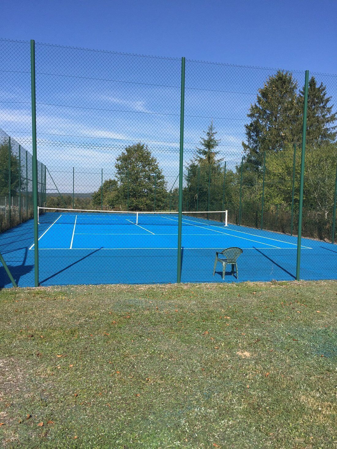 Un terrain de tennis entouré d’une clôture en treillis métallique, avec une chaise et des arbres en arrière-plan.