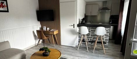 Una sala moderna con un sofá, una mesa de centro de madera y una planta en maceta. Se aprecia un comedor con una isla de piedra y sillas blancas. En el fondo, se distingue una cocina con estufa y armarios.
