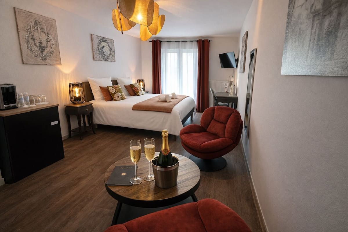 Habitación de hotel con cama, un sillón rojo, una mesita con vasos y una botella, y obras de arte en las paredes.