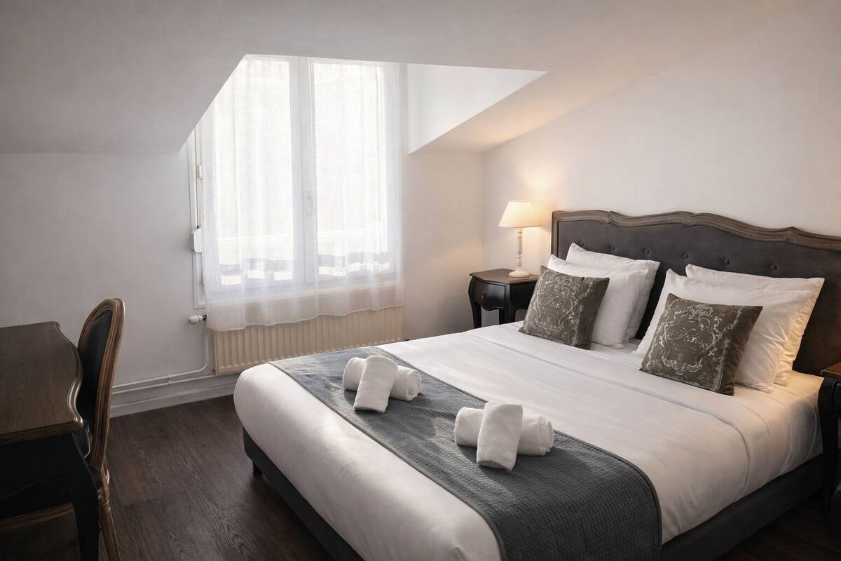 Una habitación de hotel con cama, escritorio, silla, mesita de noche, lámpara y ventana con cortinas traslúcidas.