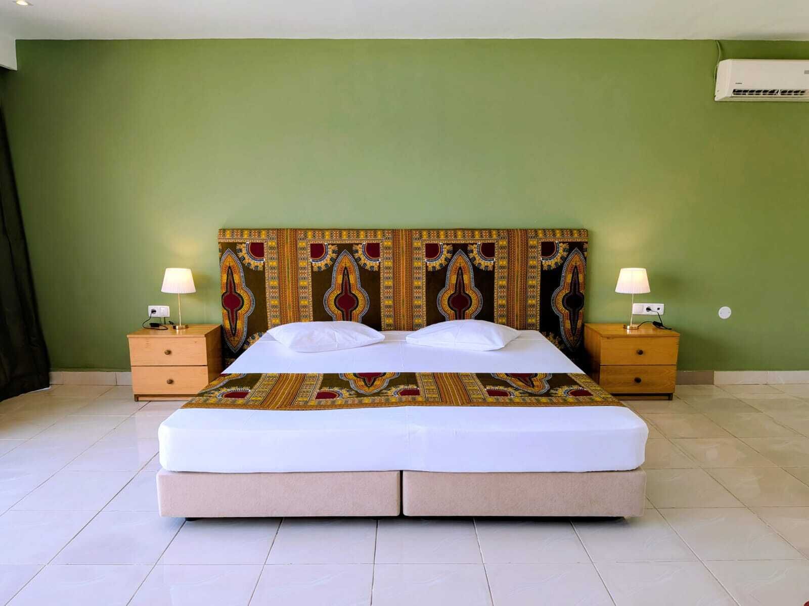 A double bed with a patterned headboard, two bedside tables with lamps, and a green wall with an air conditioning unit.
