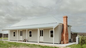 A single-story house with a white exterior, a brick chimney, and a covered porch. - Cosy 2 bedroom cottage with rural farmland views (Ross Creek)