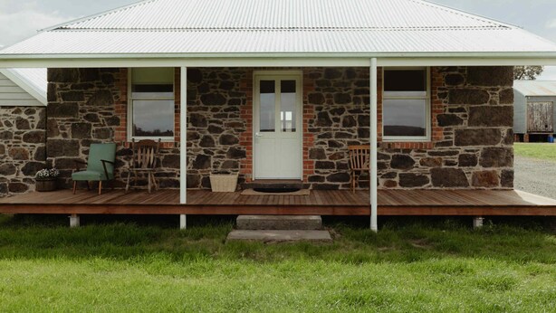A stone house with a white door and windows, a porch with chairs, and a wooden deck. - Cosy 2 bedroom cottage with rural farmland views (Ross Creek)