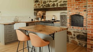 A kitchen with a stone wall, a brick fireplace, and a wooden island with two bar stools. - Cosy 2 bedroom cottage with rural farmland views (Ross Creek)