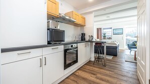 Ett modernt kök med vita skåp, en inbyggd ugn och en mikrovågsugn. Det finns en matplats med ett bord och stolar, och en utsikt över omgivningarna genom ett fönster.