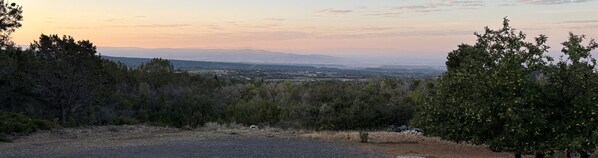 Vista panorâmica de uma paisagem rural, com uma estrada serpenteando por uma área florestal, com vista para um vale e montanhas distantes, além de uma cidade.