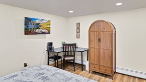Een kamer met een bed, een bureau met stoel, een houten garderobe en een schilderij aan de muur.