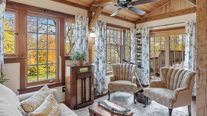 Un salon chaleureux avec des poutres en bois, un ventilateur de plafond, une grande fenêtre avec des rideaux, un canapé, un fauteuil à motifs et une table basse en bois.
