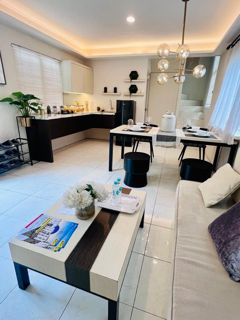 Un salon moderne comprenant un canapé, une table basse avec des magazines, un coin repas avec une table dressée pour deux personnes et un coin cuisine avec un réfrigérateur et une plante.
