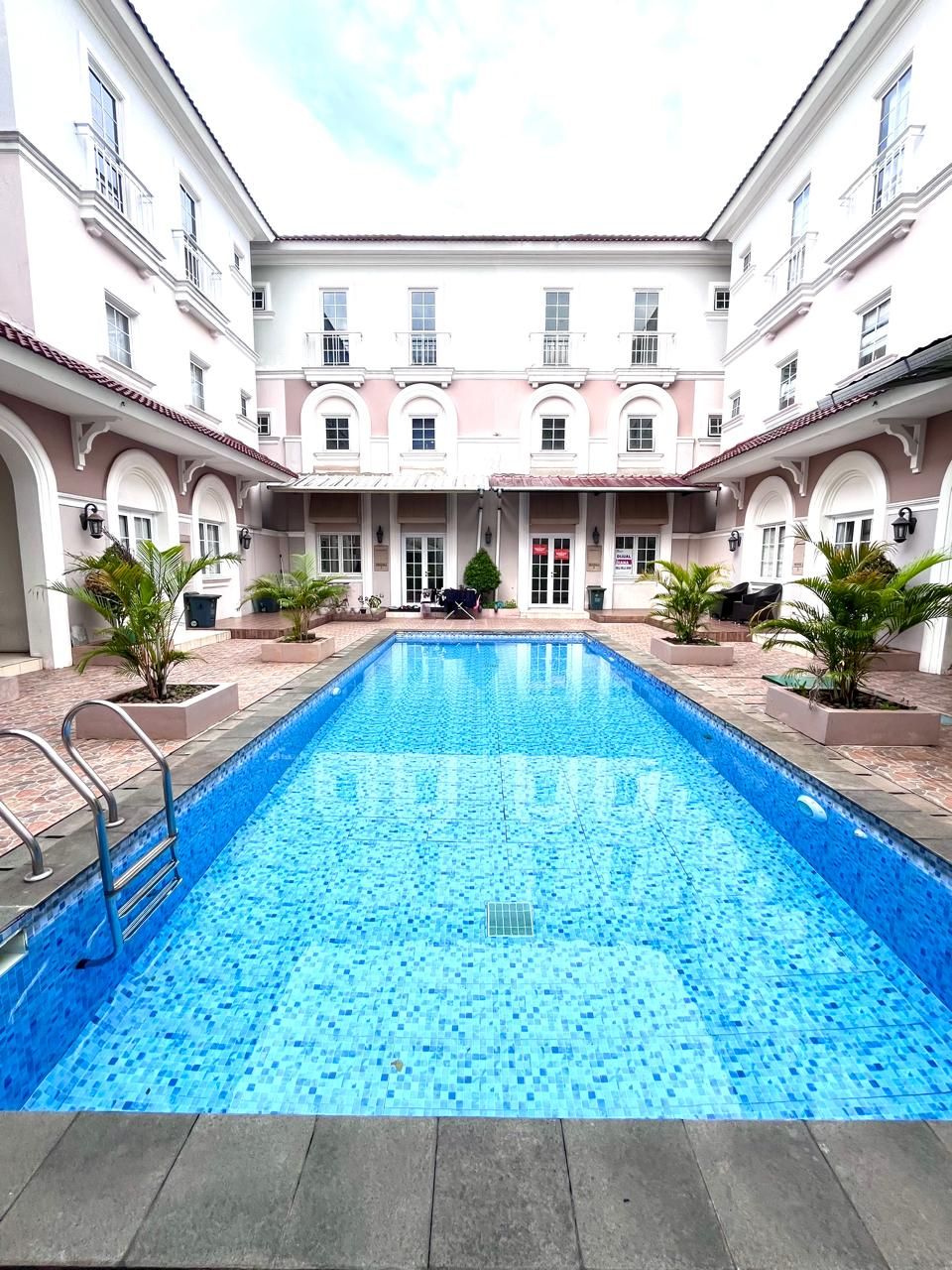 A rectangular swimming pool with a tiled edge, surrounded by a courtyard with a building featuring arched doorways and windows.