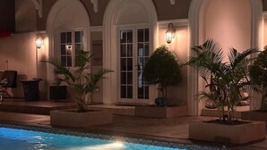 Ein Schwimmbecken mit gefliestem Beckenrand, umgeben von Kübelpflanzen und einem Gebäude mit Rundbögen und Wandleuchten.