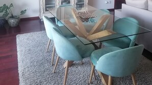 A dining area with a glass-top table, teal chairs, a white cabinet with decorative items, and a modern chandelier.