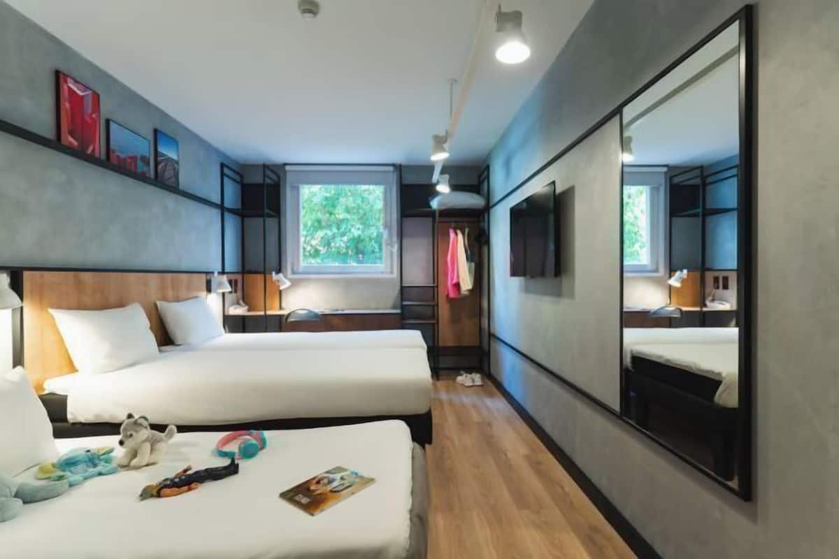 Ein Hotelzimmer mit zwei Betten, einem großen Spiegel, einem Fernseher und einem Fenster mit Blick auf Grünflächen.