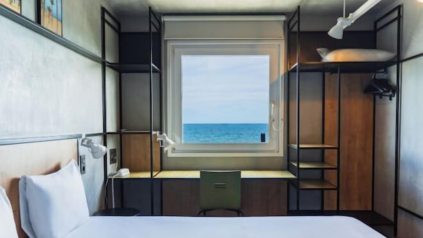 Een kleine kamer met een bed, een bureau en een raam met uitzicht op zee.