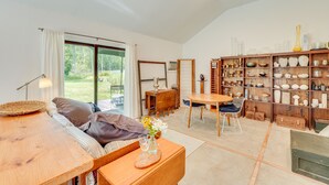 A spacious living area with a wooden table, a sofa, and a dining table. There is a large window with curtains, a bookshelf filled with decorative items, and a view of the outdoors. - 1850s Barn Studio on 4.5 Hudson Valley Acres! (Highland)