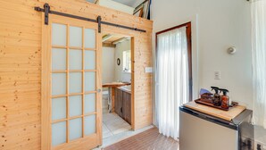 A wooden sliding door leading to a bathroom with a sink and toilet, a refrigerator, and a small table with toiletries. - 1850s Barn Studio on 4.5 Hudson Valley Acres! (Highland)