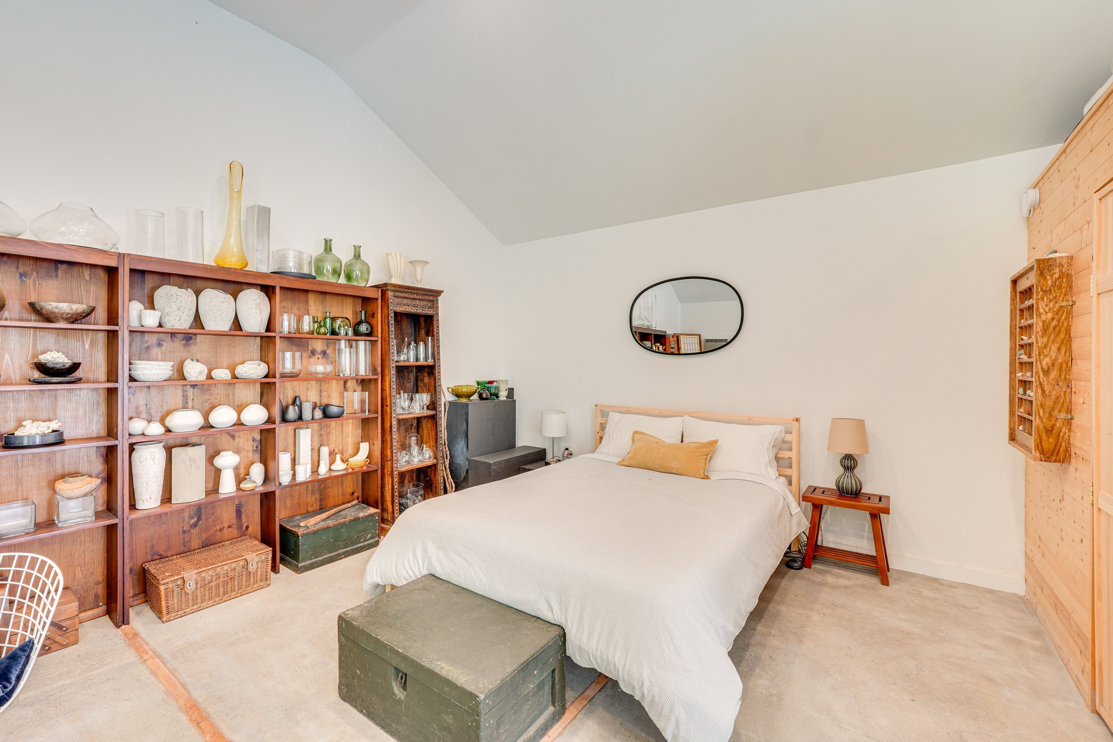 A bedroom with a large bed, a wooden wardrobe, a mirror, and shelves with various items.