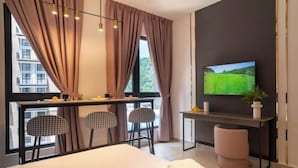 Une chambre d’hôtel comprenant un lit, un bureau avec une télévision, deux chaises, une fenêtre avec des rideaux et une vue sur les bâtiments environnants.