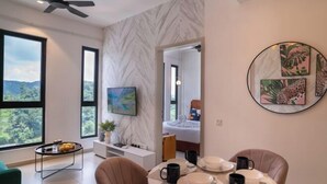 Une chambre d’hôtel moderne dotée d’une grande fenêtre, d’un ventilateur de plafond, d’un lit, d’un coin repas avec une table dressée pour deux personnes, et d’œuvres d’art accrochées aux murs.
