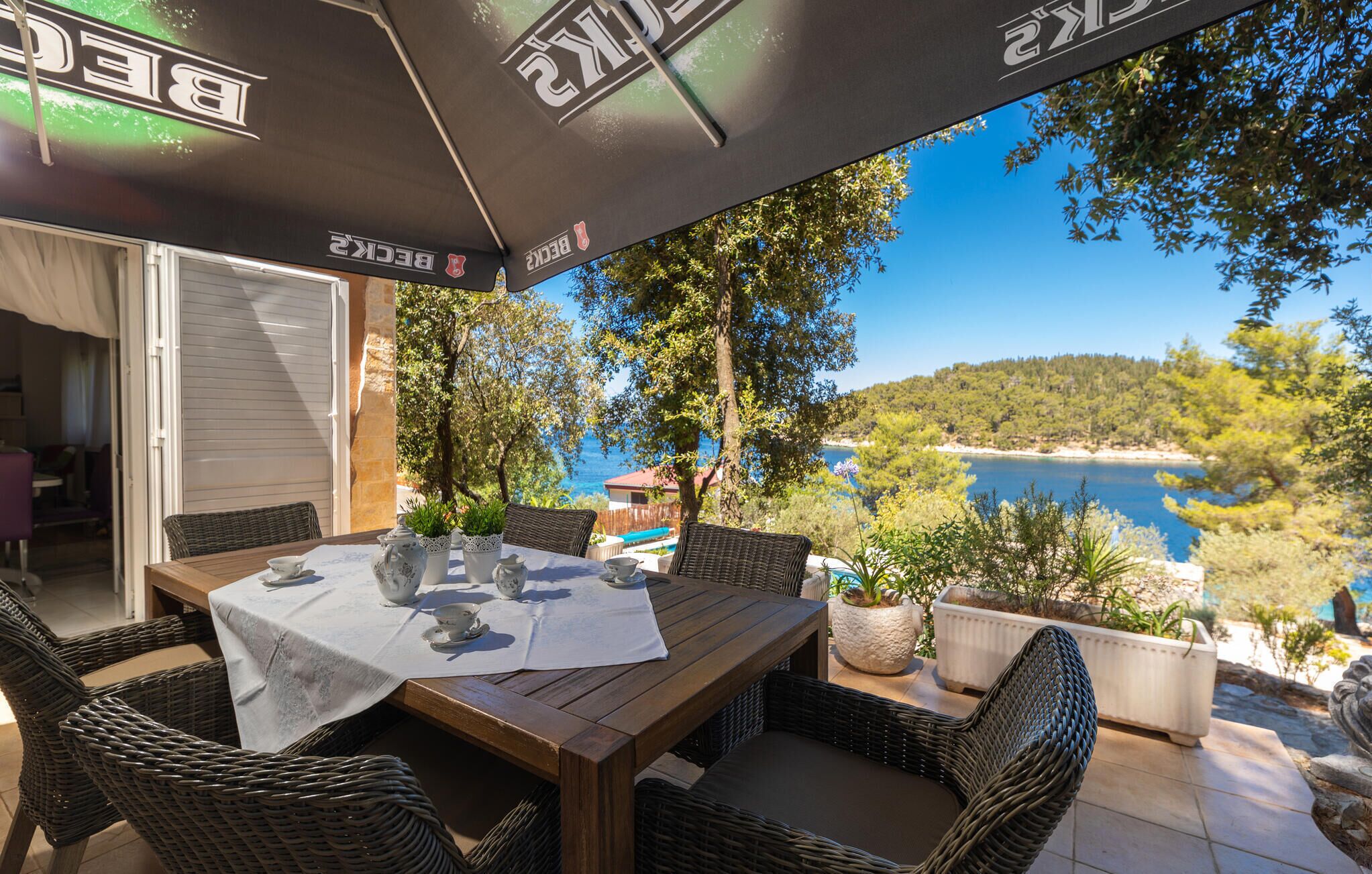 Ein Essbereich im Freien mit Korbmöbeln, einem Tisch gedeckt mit Teetassen und Blick auf Meer und Hügel.