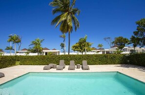 A swimming pool with lounge chairs and palm trees, surrounded by buildings with a clear blue sky. - Modern 3BR Villa-Private Pool -Sosúa Ocean Village (Sosúa)