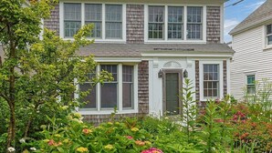 Une maison à deux étages avec un porche à l’avant, une porte et un petit jardin agrémenté de diverses plantes et fleurs.