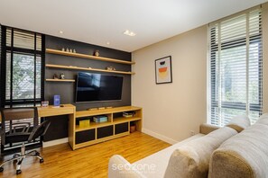 Ett modernt vardagsrum med en platt-TV, trähyllor, en soffa och ett skrivbord med en stol.