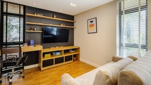 A modern living room with a flat-screen TV, wooden shelves, a sofa, and a desk with a chair. - Condominium With Excellent Location in Asa Norte - Amzs (Brasília)