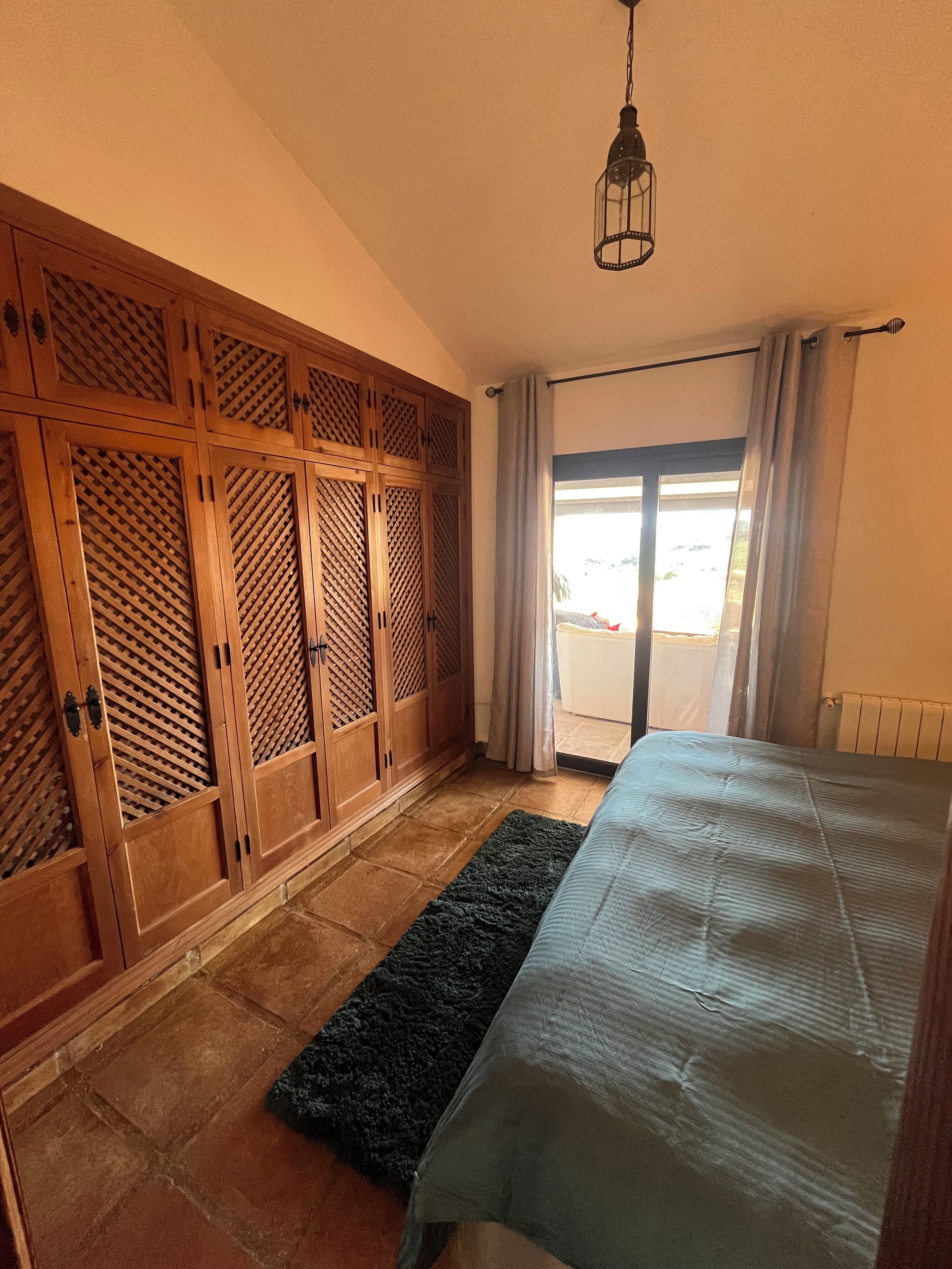 Un dormitorio con una cama, un armario de madera, una alfombra negra y una lámpara colgante.