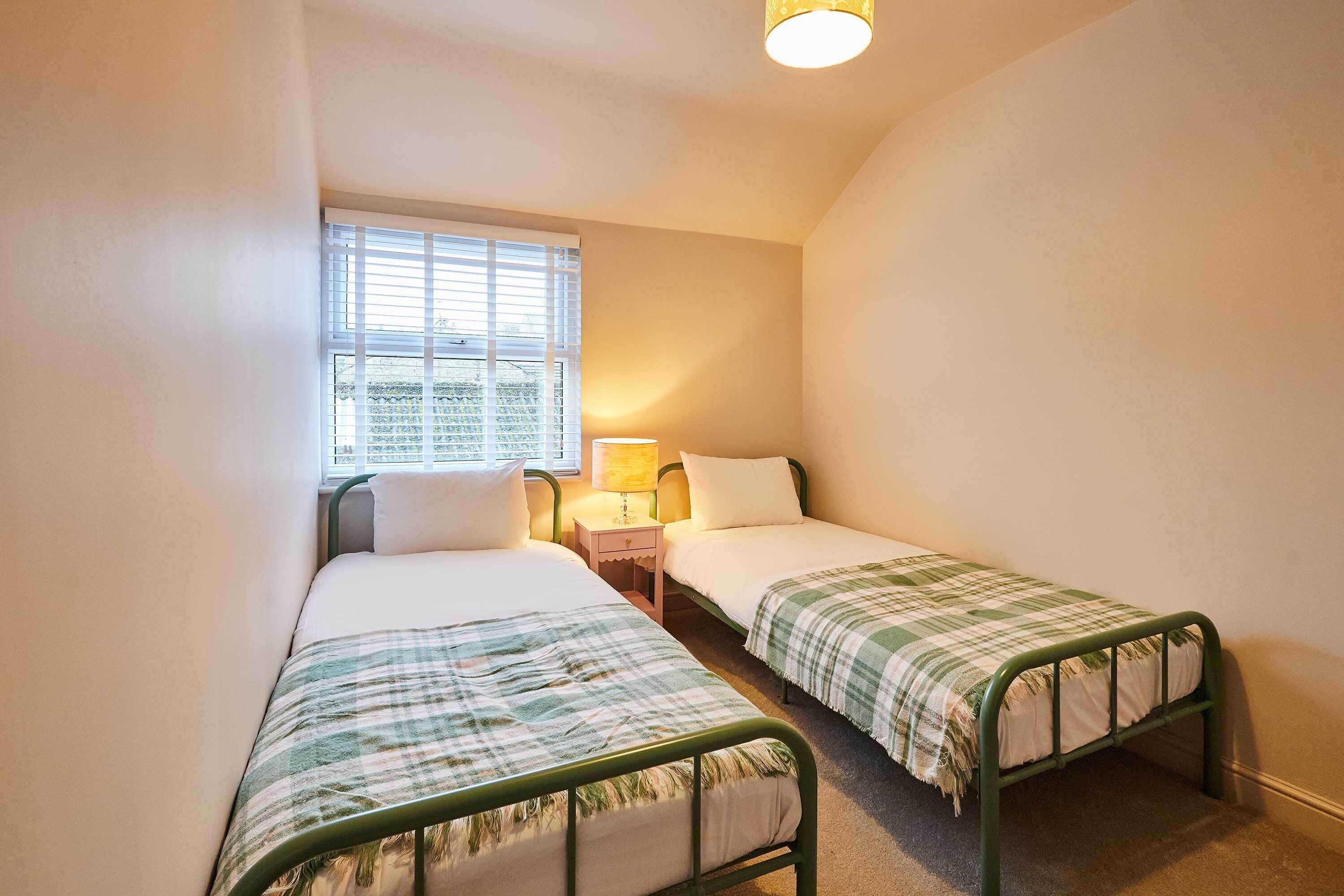 A small, single-room accommodation with two single beds, a window with blinds, a bedside table with a lamp, and a carpeted floor.