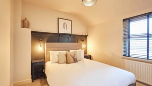 A bedroom with a large bed, bedside tables, a headboard with built-in lighting, a framed picture on the wall, and a window with blinds. - Host Stay Daisy Cottage (Ripon)