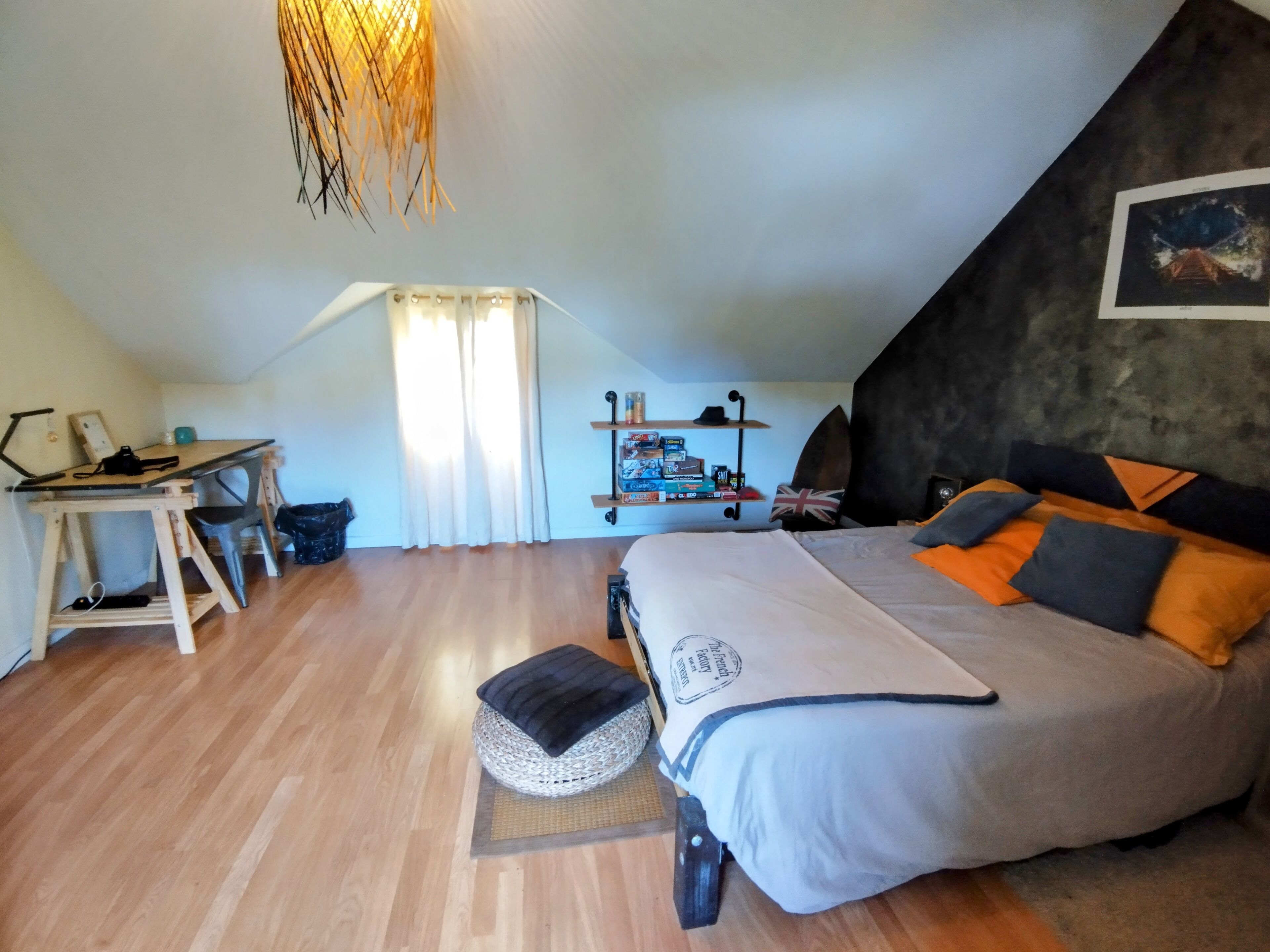 A bedroom with a bed, a desk, and a chair. There is a shelf with books and a hat.