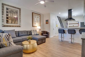 Ein modernes Wohnzimmer mit einem grauen Sofa, einem runden Couchtisch und einer Treppe, die in einen Küchenbereich mit Barhocker führt.