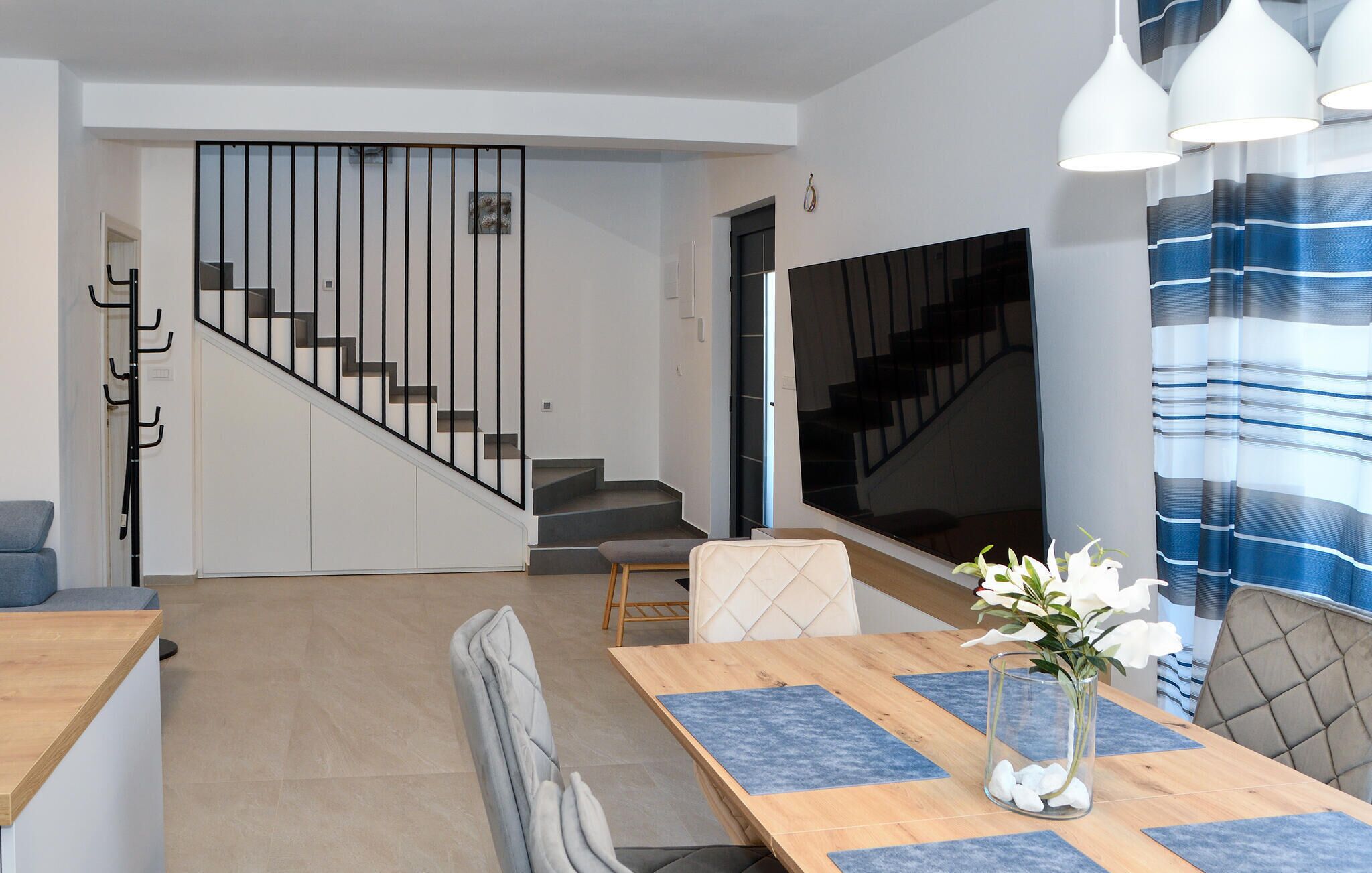 A modern living room with a staircase, a dining area with a table and chairs, and a large window with curtains.