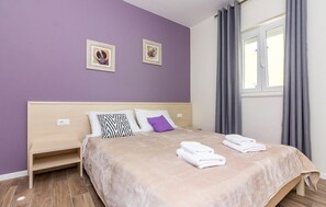 Una cama bien hecha con una colcha color beige y dos almohadas, una mesita de noche de madera y una ventana con cortinas grises.