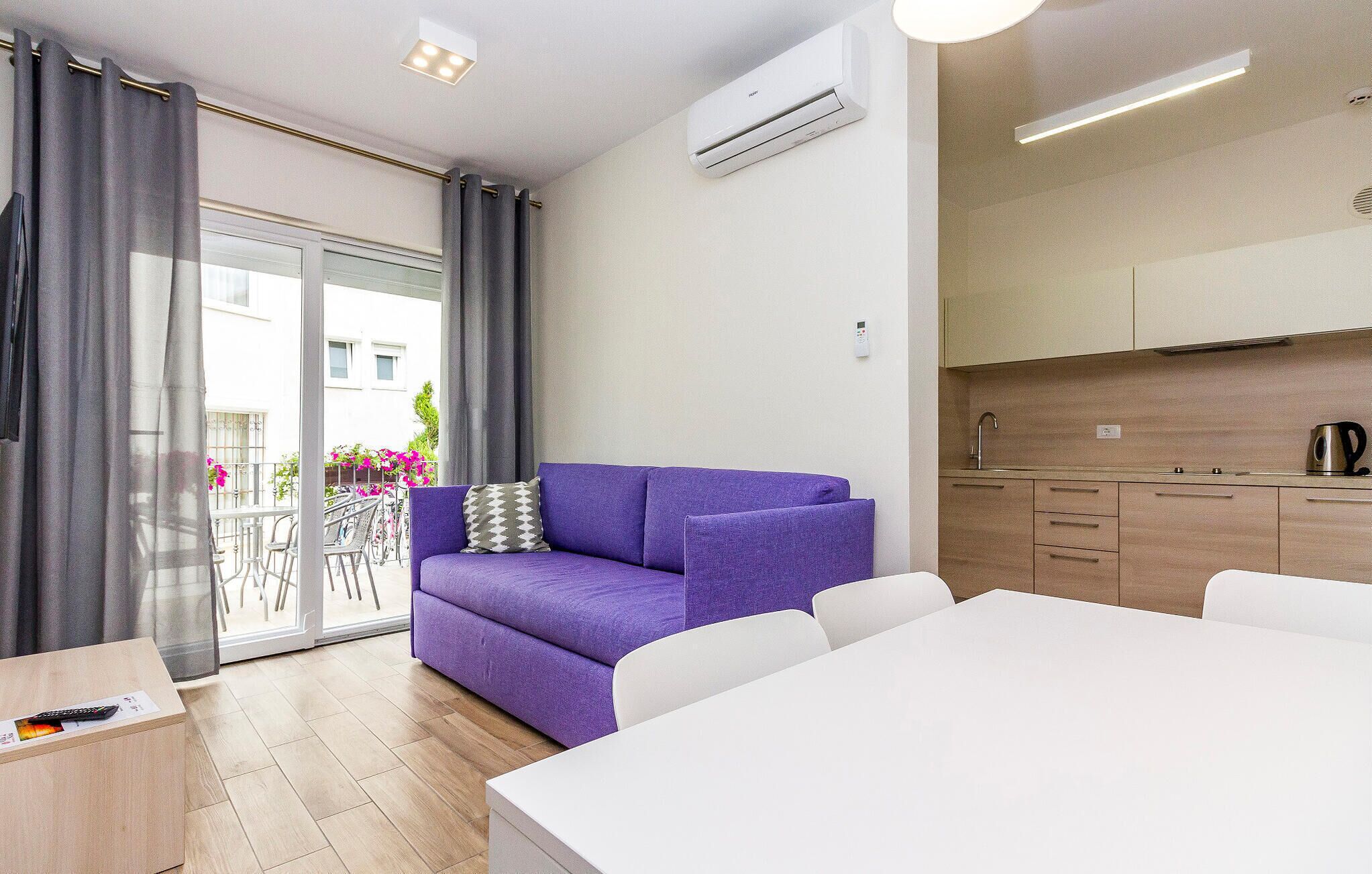 A modern living room with a purple sofa, a white dining table, and a kitchen area with wooden cabinets.