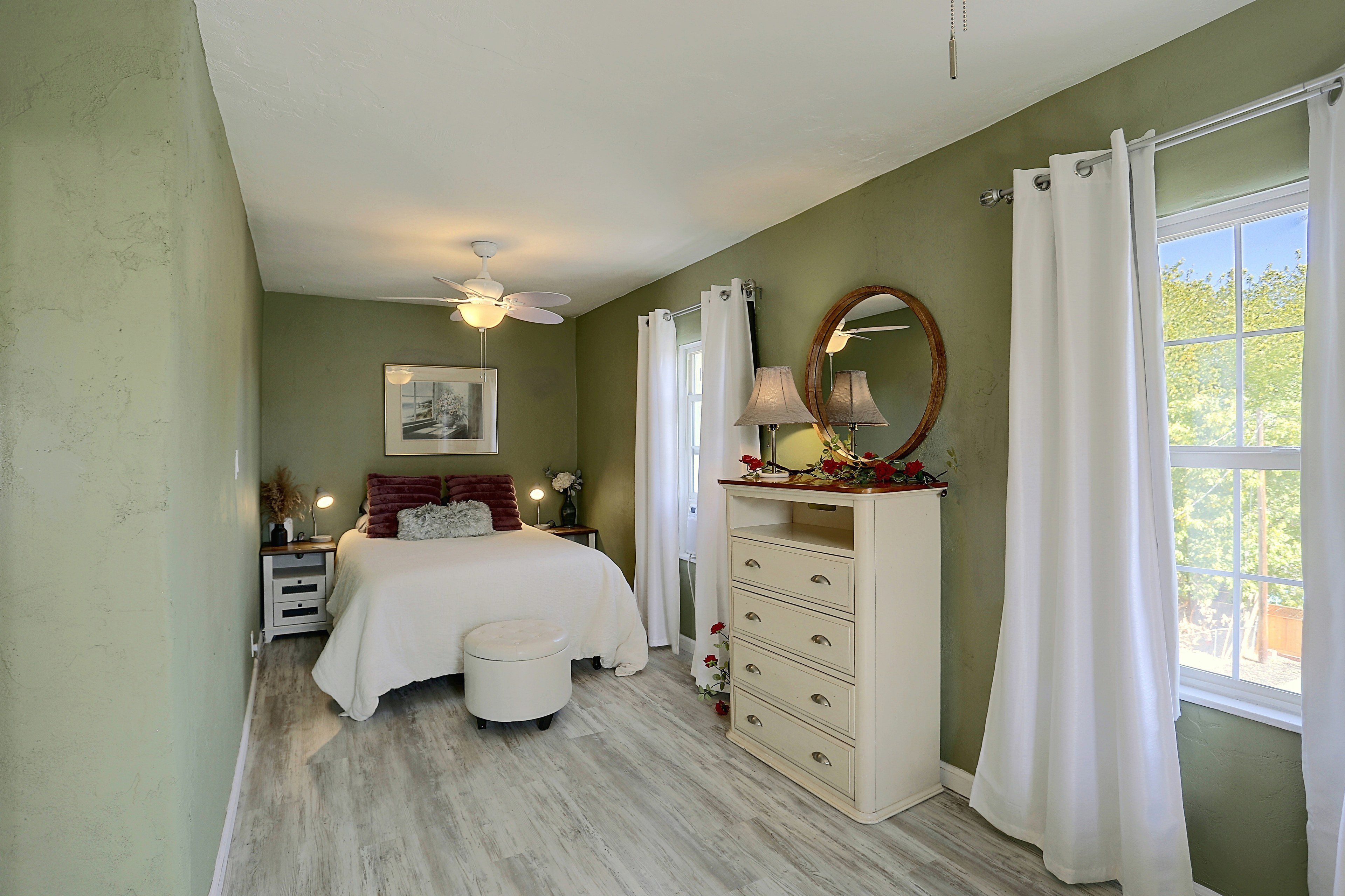 Un dormitorio con cama, mesitas de noche, cómoda, espejo y ventana con cortinas.