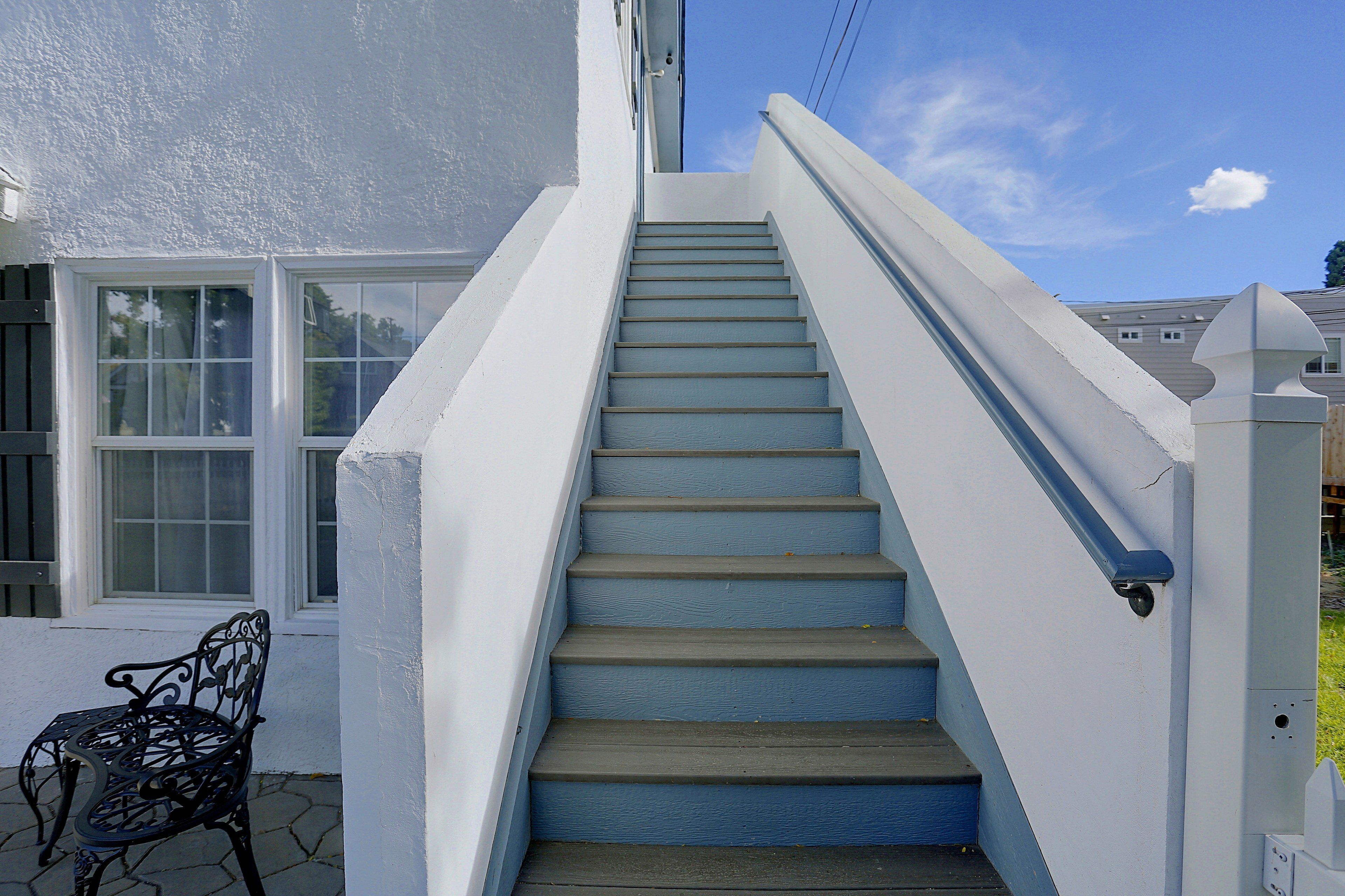 A set of blue-painted exterior stairs leading to a white building with a window and a metal bench.