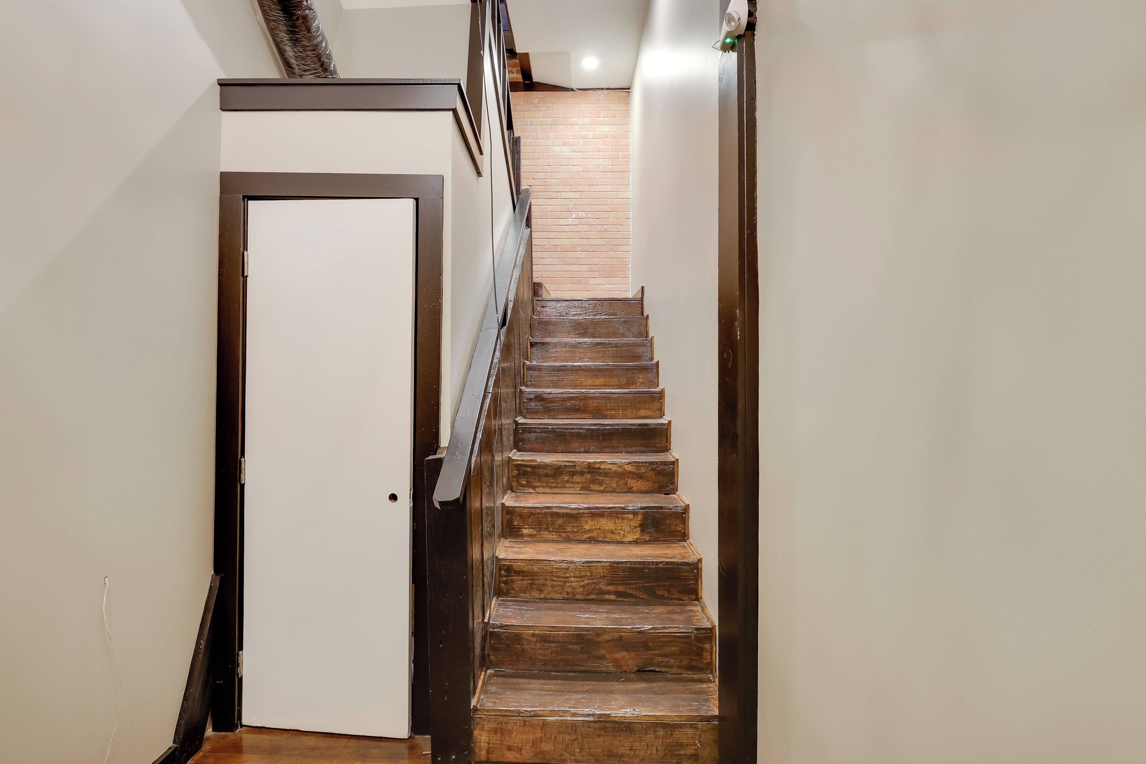 Un escalier en bois, avec une porte blanche en haut, mène à une pièce dont un des murs est en briques.