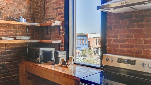 A kitchen with a brick wall, a window, a microwave, a stove, and shelves with dishes. - Convenient Couple's Retreat in Dtwn Marshall! (Marshall)