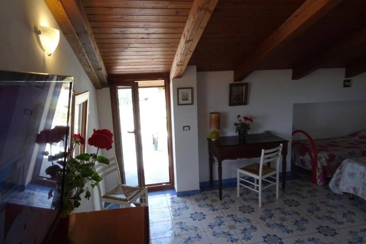 A room with a wooden ceiling, a bed with a floral bedspread, a small table with a chair, and a door leading outside.
